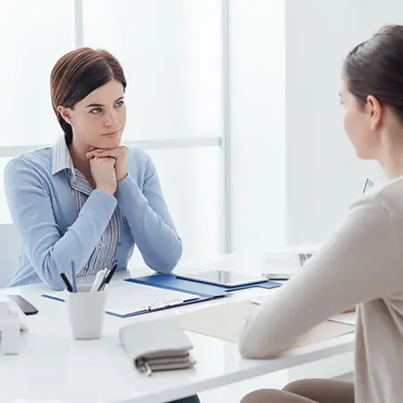 Two office workers sit across a table in a one-on-one meeting in a corporate office.